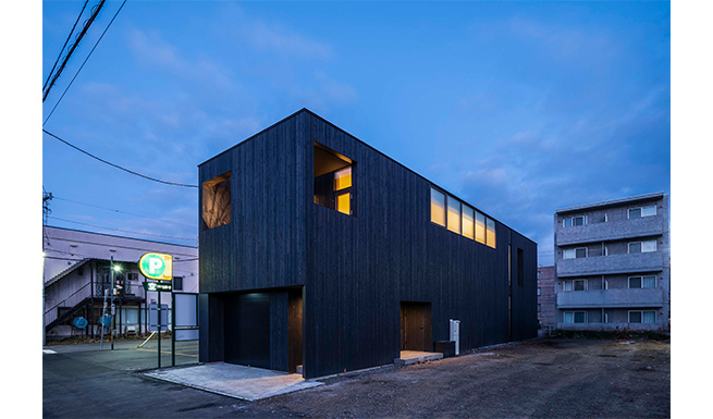 南東外観夕景。両隣は駐車場で開けており、建物を見通せる。